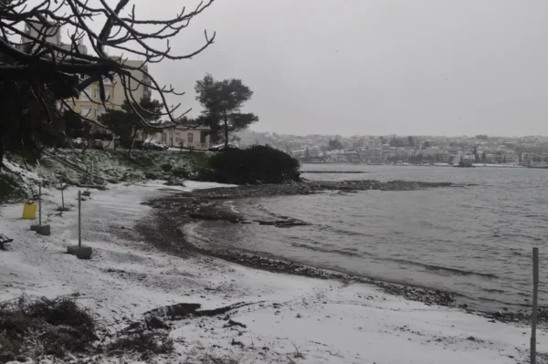 Snow blankets the pebble beach and shoreline of Chalkis, Euboea, while trees and a distant town peek through the winter scene.