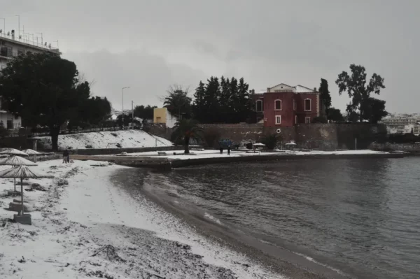 Chalkis features a snowy beach scene with a calm sea, palm trees, and a historic pink building along the shoreline in Euboea, Greece.