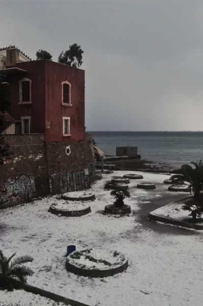 Snow-covered coastline of Chalkis, Euboea, Greece, featuring a pink building, stone walls, and landscaped circular areas.