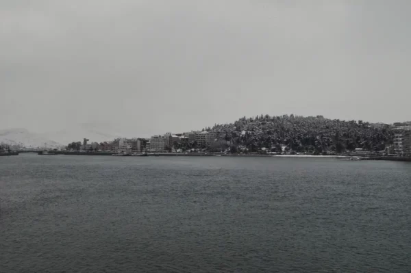 Snow-covered hills rise above a serene harbor in Chalkis, Euboea, Greece, framed by muted gray skies and tranquil waters.