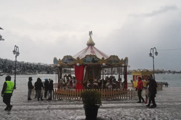 Colorful carousel with ornate details stands by the snowy waterfront in Chalkis, Euboea, Greece, with visitors exploring nearby.