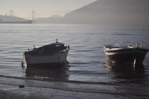 Two boats gently float on calm waters in Chalkis, Euboea, Greece, with a hazy backdrop of hills and a distant bridge.