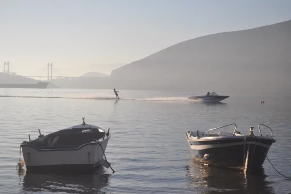 Boats in calm waters near Chalkis, Euboea, Greece, with a wakeboarder gliding past against a misty backdrop.