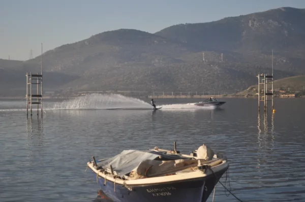 Skiers glide across the calm waters near Chalkis, Euboea, Greece, with scenic mountains in the background and a boat in the foreground.