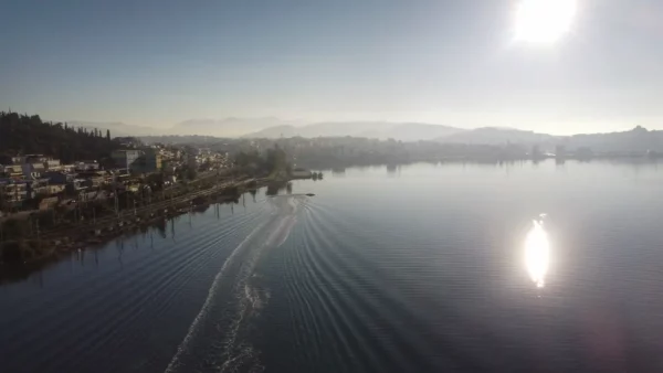 Calm waters reflect sunlight near Chalkis, Euboea, with a shoreline of trees and buildings against a misty mountain backdrop.
