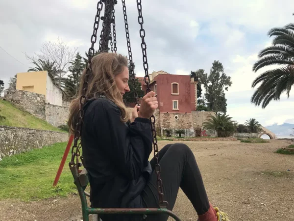 A woman sits on a swing in Chalkis, Euboea, Greece, with palm trees and historic buildings in the background under a cloudy sky.
