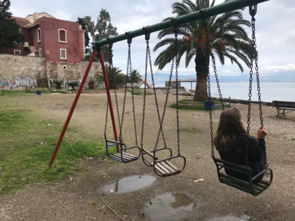 Empty swings in a park overlooking the sea in Chalkis, Euboea, Greece, framed by palm trees and a historic building.