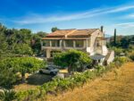 Sunlit hillside property with a white house and red-tile roof overlooking greenery in Corfu, Greece