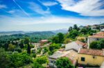 Wide view from a hilltop overlooking Corfu, Greece, with houses, trees, and distant blue mountains under a partly cloudy sky.