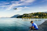 Man fishing from a dock over calm water, with a bridge and hillside buildings along the shoreline under a blue sky.