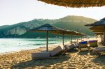 Beachside lounge chairs under umbrellas with calm sea and green hills in the distance, Corfu, Greece