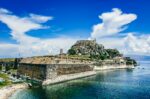 Old fortress in Corfu Town, Greece, with stone ramparts and a small harbor along the waterfront under a blue sky with clouds