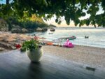 Corfu, Greece beach scene with potted flowers on a terrace overlooking calm water, boats moored near the shore, and umbrellas on the sand.