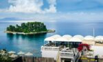Corfu town Greece in 2026, overlooking the sea from a seaside terrace with white domes, a pool, and a small green island in the distance